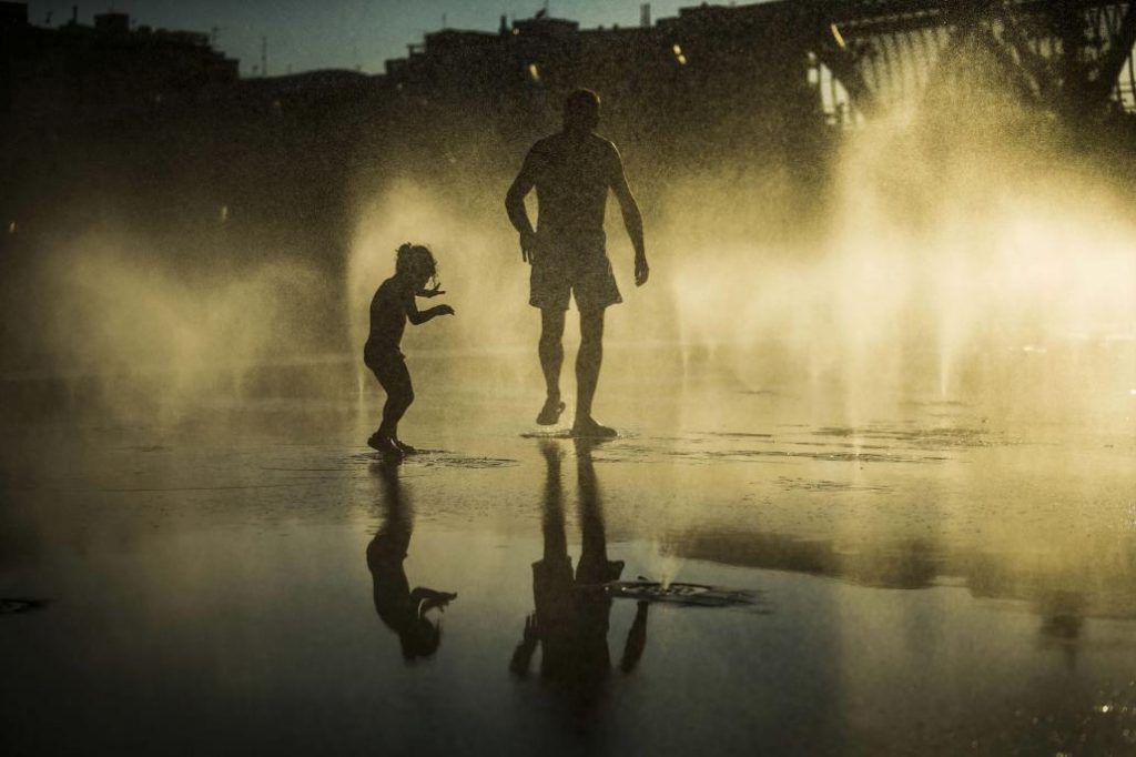 Olas de calor. Un hombre y una niña tratan de combatir la ola de calor refrescándose en una fuente de surtidores vaporizados